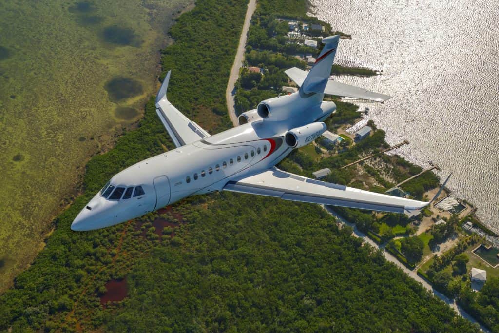 Falcon 900LX aircraft flying over a coastal area with lush greenery and water, featuring red and blue accents on the tail. 2